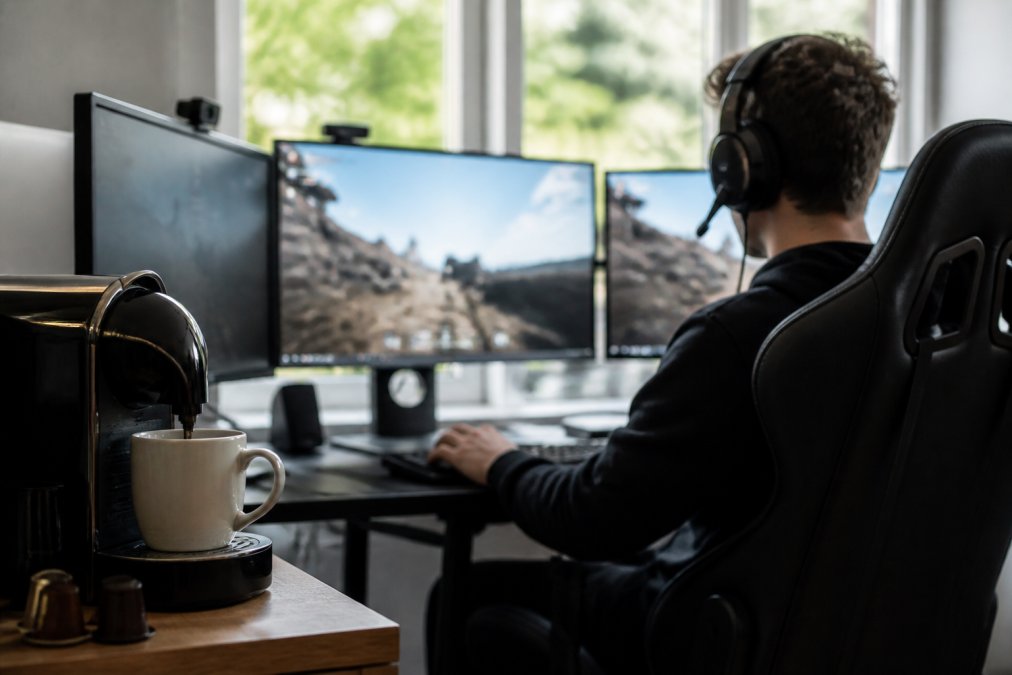 Gamer seated at desk with multiple monitors displaying competitive game interface, coffee pod brewing station with mug in foreground, focused expression during active gameplay, natural window lighting