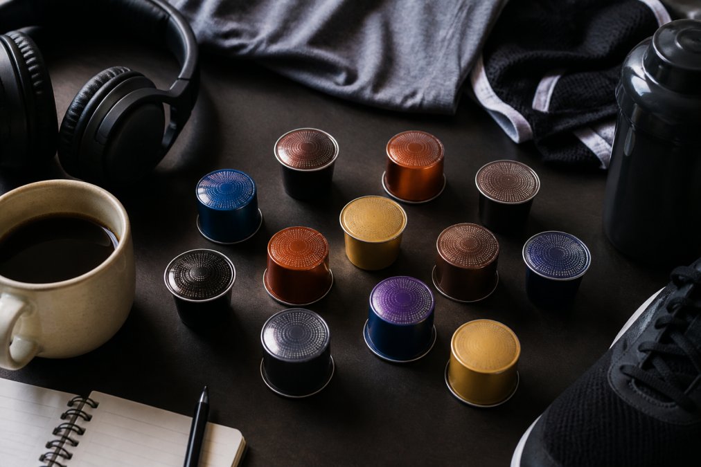 Overhead flat lay arrangement of coffee pods displayed on desk with ceramic mug, gaming headphones, notebook, and athletic gear visible in background, natural office lighting