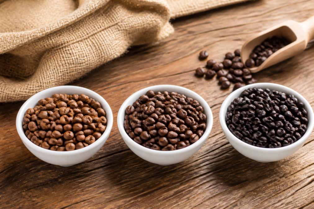 Three small white bowls on a wooden table, each filled with coffee beans of different roast levels: light brown, medium brown, and dark almost black, with a burlap sack and a wooden scoop nearby