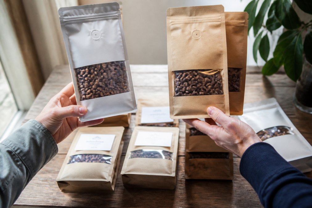 Close-up of hands holding different coffee bags with visible roast dates and tasting notes labels, displaying beans with varying roast colors in natural window light