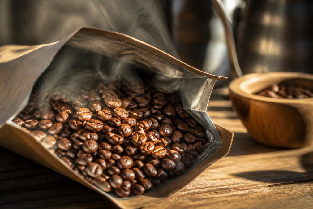 Close-up of freshly roasted coffee beans in a sealed kraft paper bag with visible steam rising, afternoon sunlight highlighting the glossy bean surface and roasted color variation