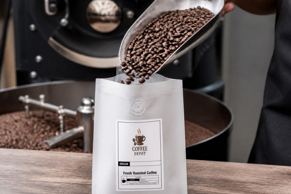 Close-up of freshly roasted dark brown coffee beans being carefully poured into a white vacuum-sealed bag with a one-way valve, industrial roasting equipment visible in the soft background