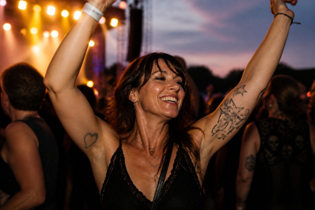 A woman in her forties with a big grin, dancing with friends at an outdoor live music gig at dusk, hands in the air, warm stage lights behind her