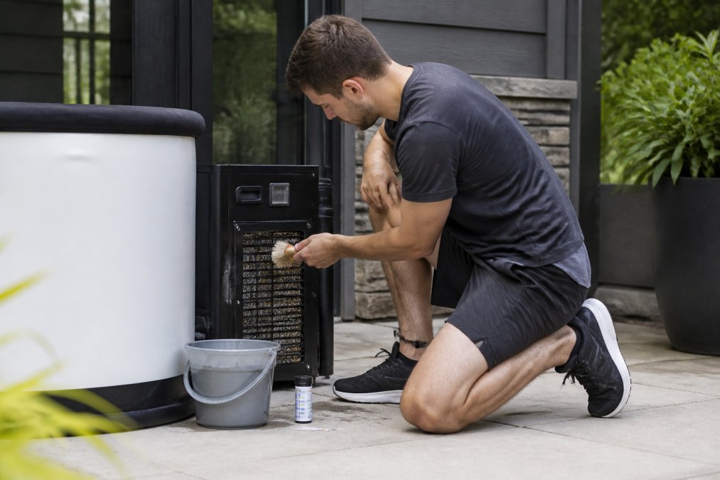 A person in casual workout clothes kneeling beside a modern cold plunge tub on a home patio, wiping the chiller's condenser coils with a soft brush, with a small bucket and test strips nearby