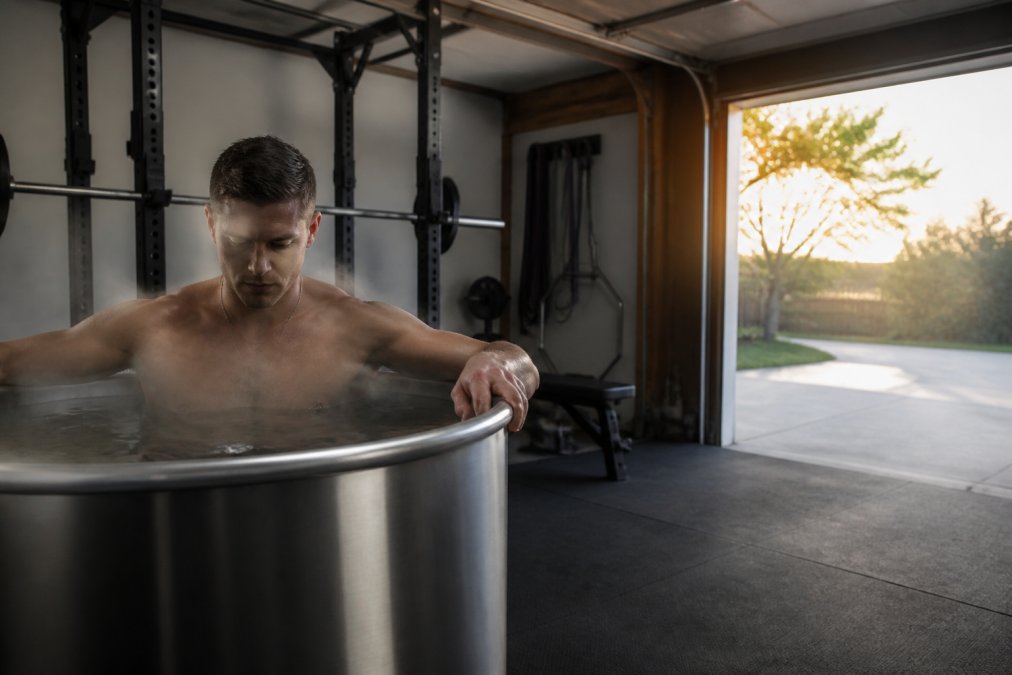 Strength athlete stepping into a stainless steel cold plunge in a morning garage gym setting