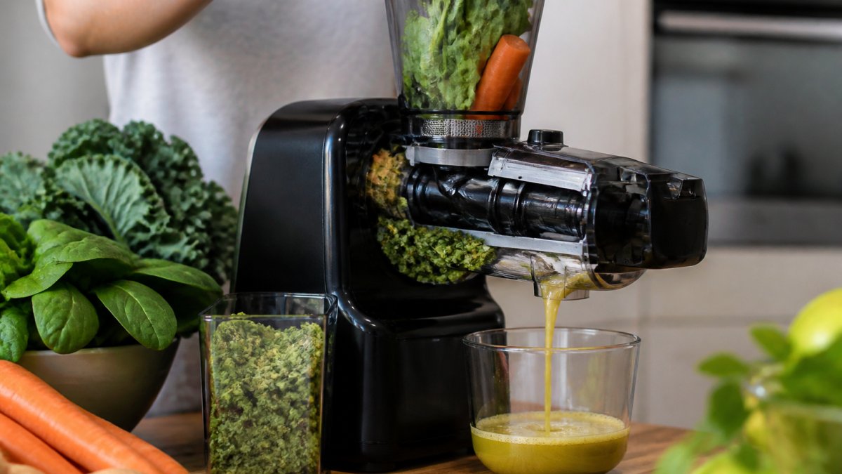 Close-up of fresh produce including kale, spinach, and carrots being fed into a cold-pressed juicer with visible auger mechanism and golden juice flowing into a glass container