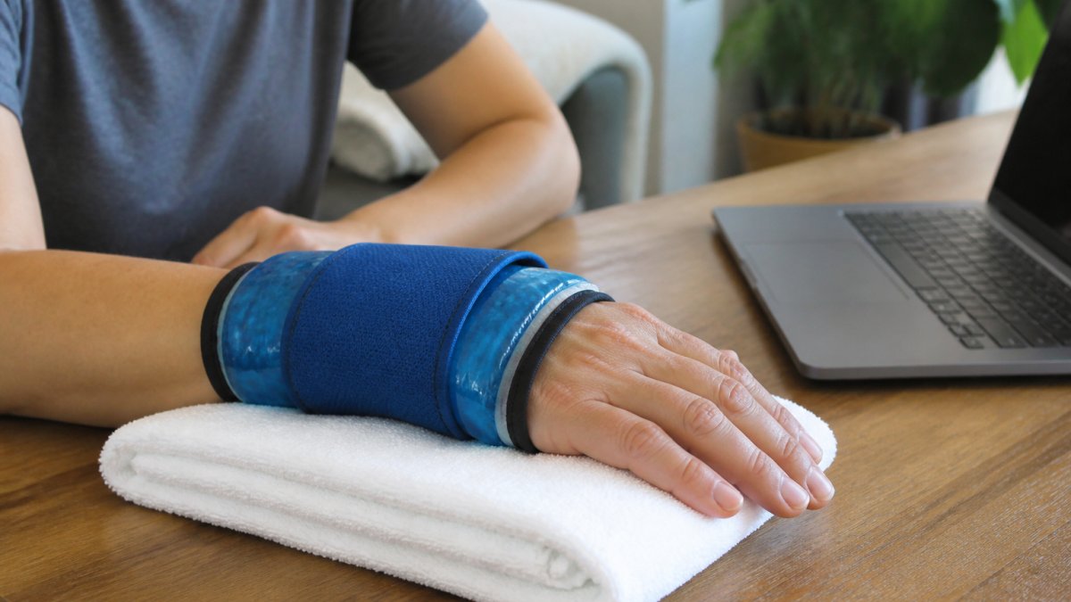 A close-up photo of a person's hand and wrist wearing a blue gel cold compression wrap while resting on a table, with a towel underneath and a laptop nearby, showing a calm home setting