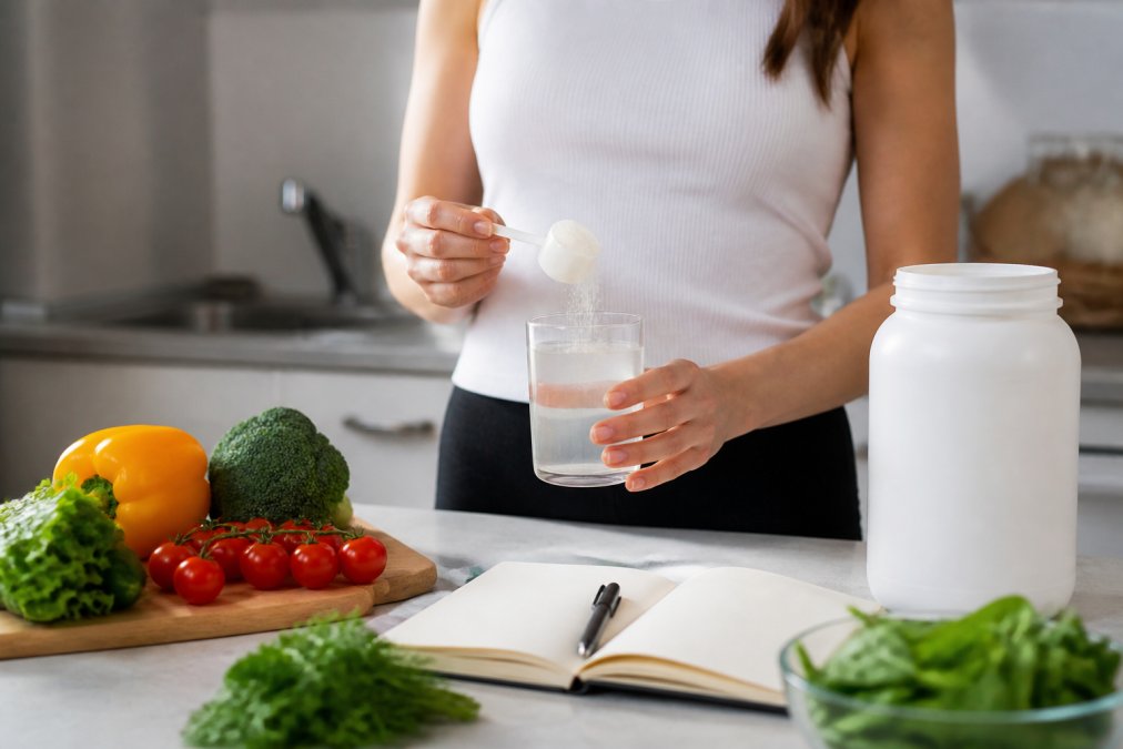 Close-up of a person holding a glass of collagen peptide powder mixed with water, with fresh vegetables and a wellness journal on a clean kitchen counter