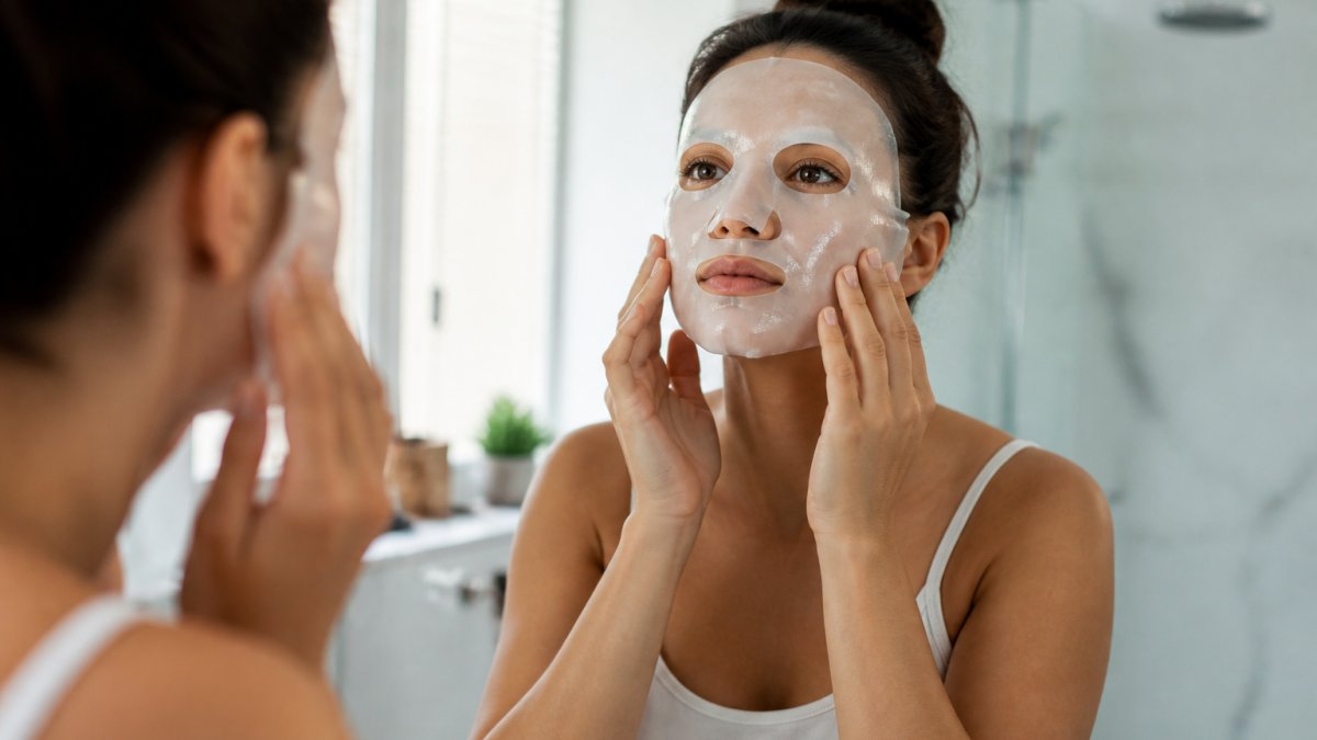 Woman applying a hydrogel collagen mask to her face in a bright, clean bathroom with soft lighting, showing proper placement and technique