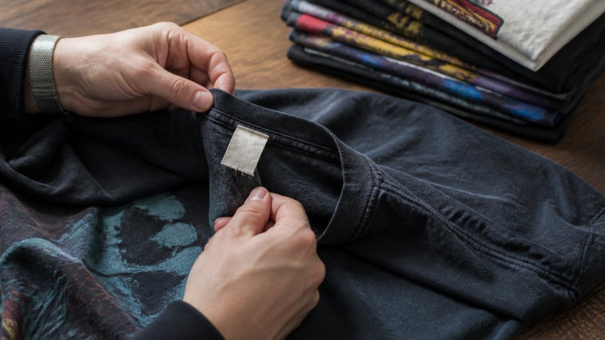 Close-up of hands examining the hem and printed tag of a faded vintage graphic tee laid flat on a wooden table, with a stack of retro tees in the background