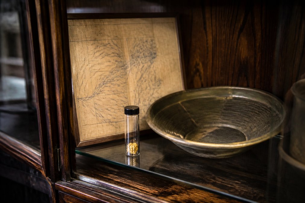A well-organized display case featuring a raw placer gold flake specimen in a small glass vial, an antique gold pan, and a vintage mining claim map, set against a dark wooden backdrop with warm ambient lighting