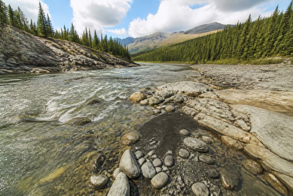 Wide shot of a rocky riverbed with visible bedrock formations, bends, and areas where black sands accumulate along the water's edge in clear daylight