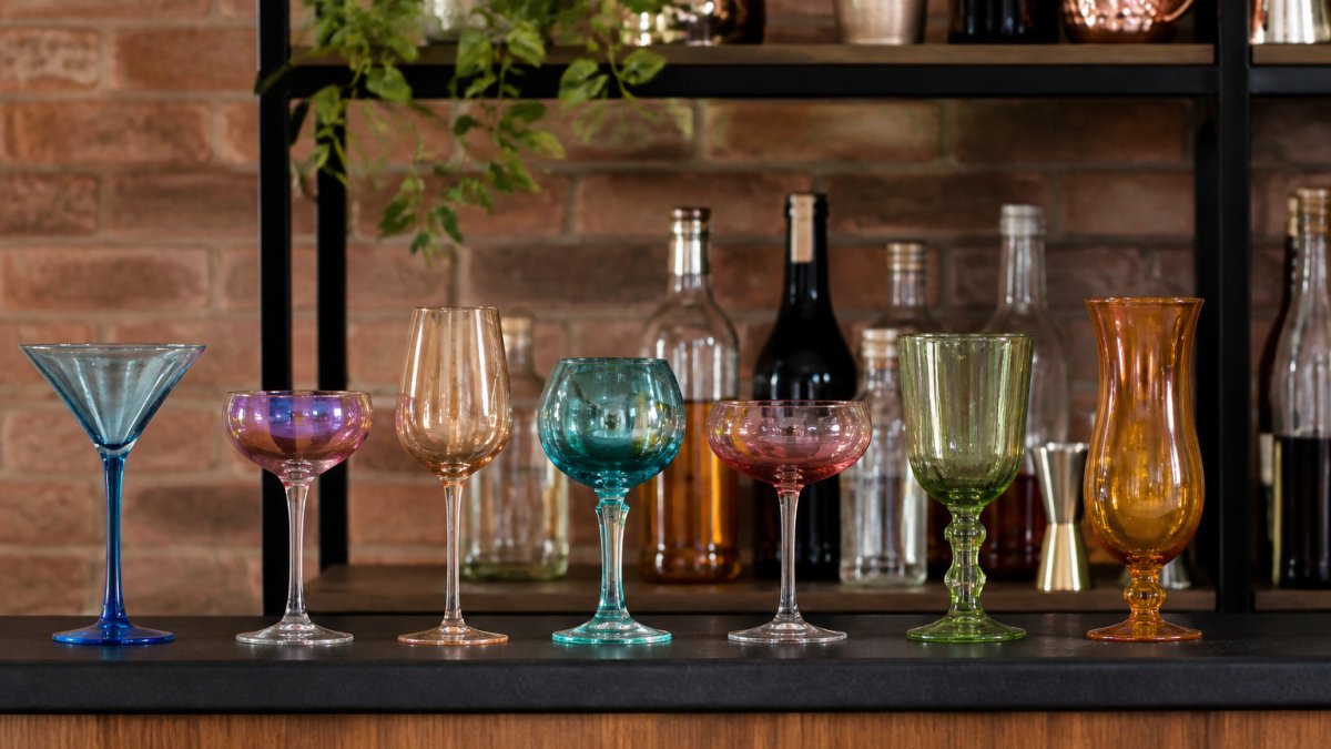 Home bar setup featuring an assortment of colorful cocktail glasses in different styles and hues, martini, coupe, wine, and hurricane glasses, arranged on a shelf with bottles and bar tools in the background