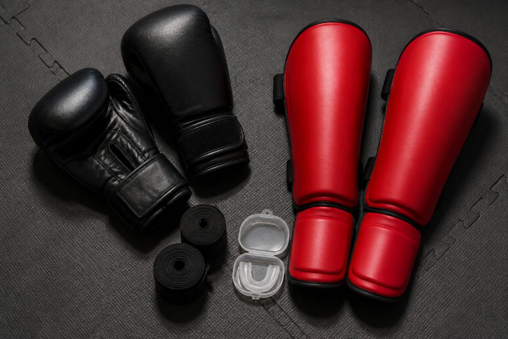 A flat lay of essential beginner combat gear: black boxing gloves, rolled hand wraps, a clear mouthguard case, and red shin guards arranged neatly on a dark gym floor with natural overhead lighting