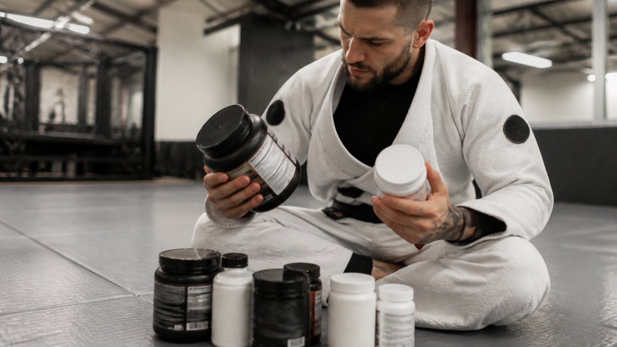 Grappler in gi examining supplement bottles and reading ingredient labels in a bright training facility, carefully studying dosage information on multiple containers