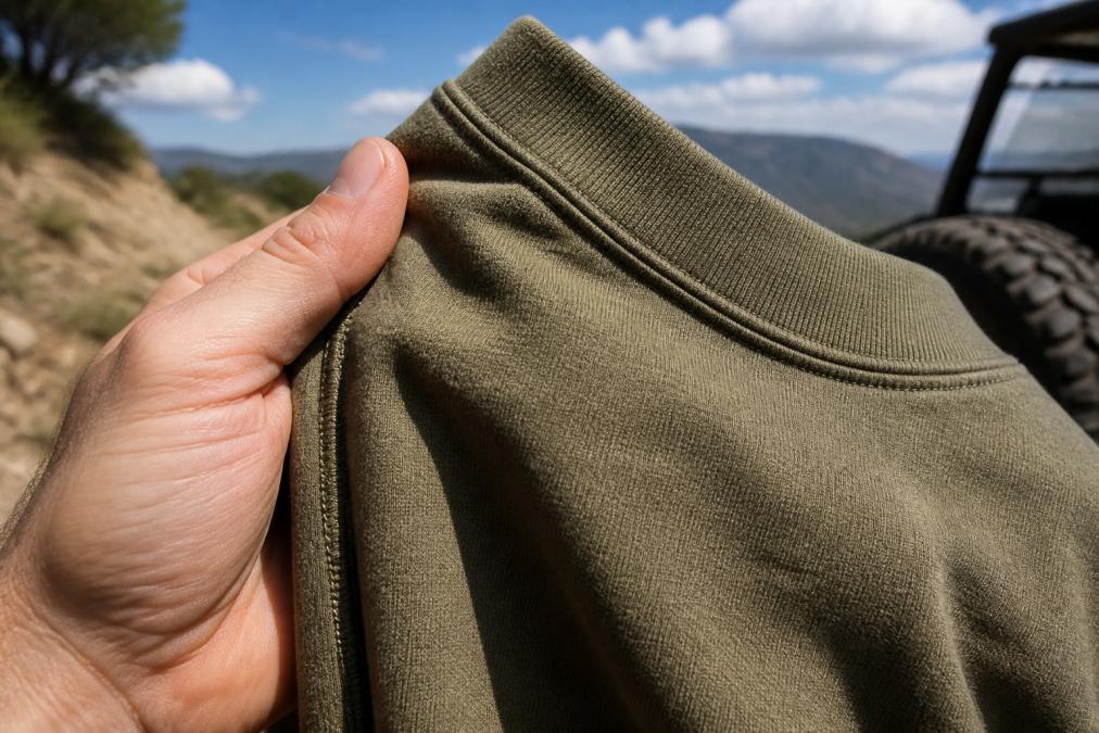 Close-up of a hand gripping the thick, textured fabric of a heavyweight t-shirt, showing the ribbed collar and side-seam stitching in a sunny outdoor setting