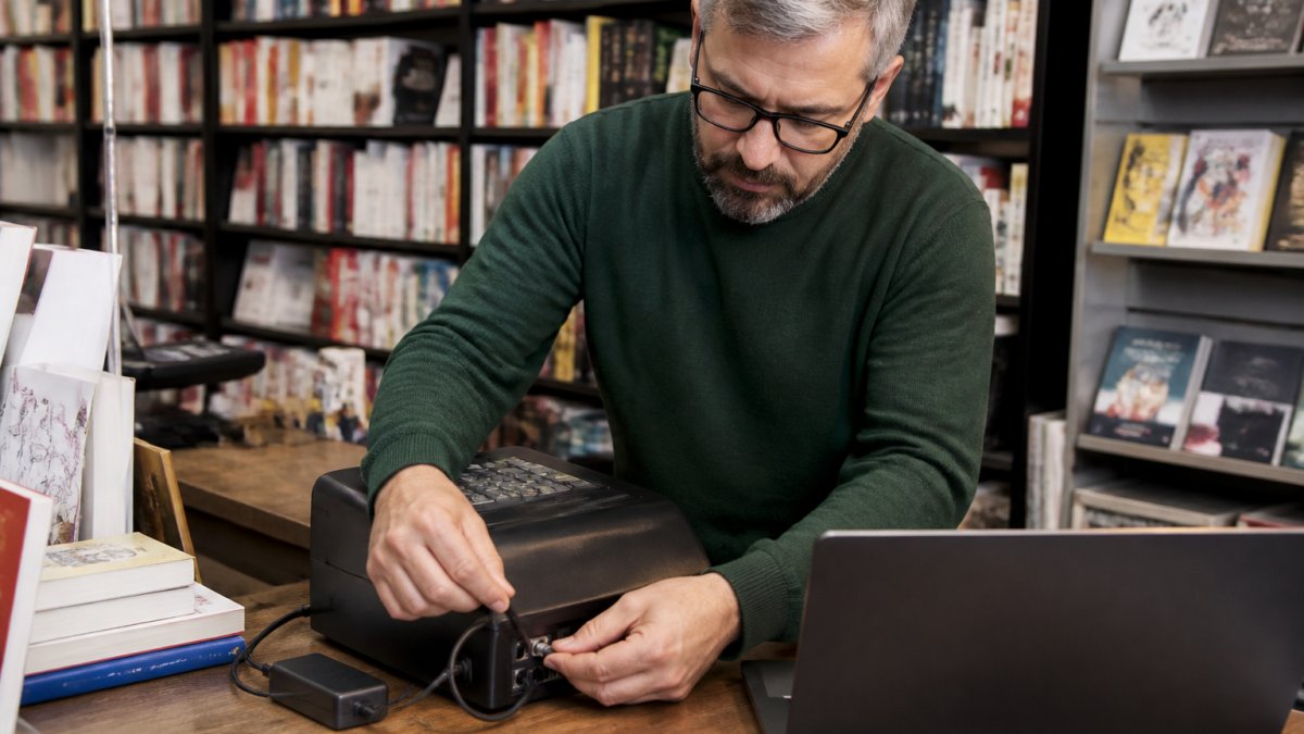 Un libraio che configura un registratore telematico accanto a un computer portatile sul bancone di una libreria indipendente, con scaffali di libri sullo sfondo