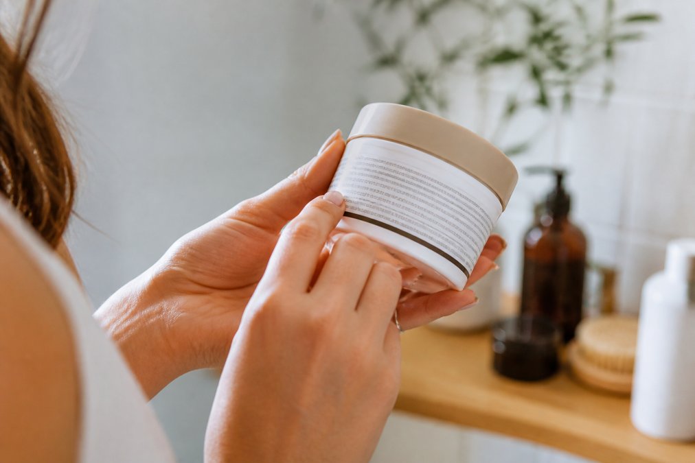 Mani femminili che tengono un barattolo di crema e lo girano per leggere la lista degli ingredienti sull'etichetta, luce naturale soffusa