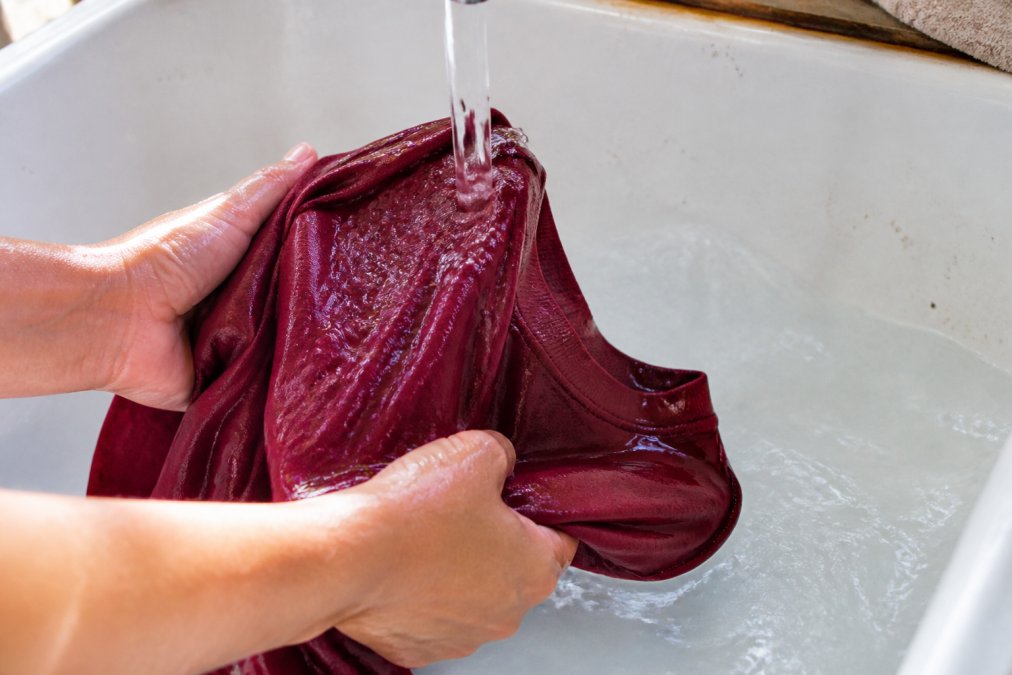 Primo piano di una maglietta di cotone appena tinta in colore ricco che viene risciacquata sotto acqua corrente cristallina, mostrando l'ultimo passaggio della fissazione del colore