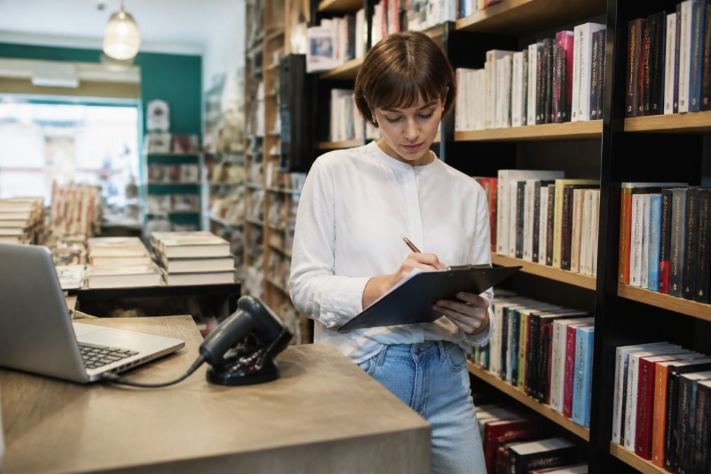 A bookseller in a small independent bookstore writing inventory notes on a clipboard next to a shelf of books, with a laptop and barcode scanner on the counter nearby