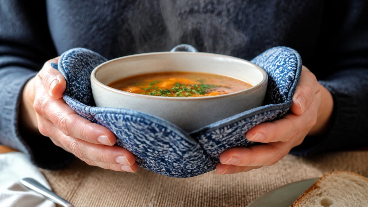 Close-up of hands holding a steaming hot ceramic bowl with a patterned fabric cozy, showing secure grip and comfortable mealtime experience without visible heat stress