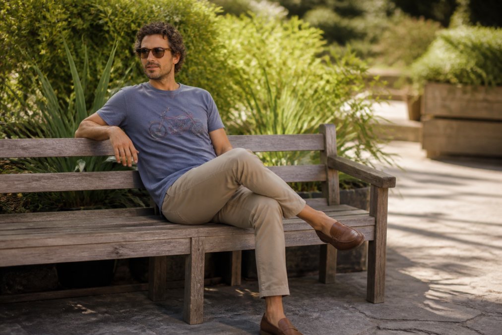 A relaxed man in his 30s wearing a soft vintage-inspired graphic tee, well-fitted chinos, and loafers, sitting casually on a timber bench outdoors on a sunny afternoon