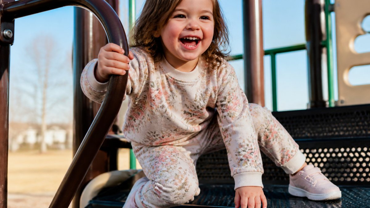 A toddler girl in a soft floral jogger set laughing while climbing on a playground structure, bright natural daylight, candid action shot
