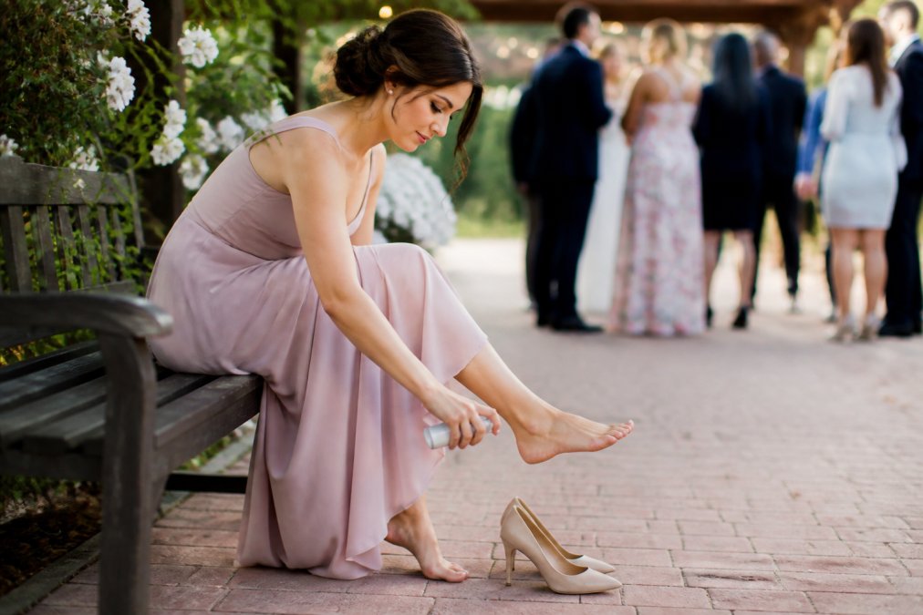 A woman sitting on a bench at an outdoor wedding venue, applying a small spray bottle to her feet before putting on her high heels, with other guests mingling in the background