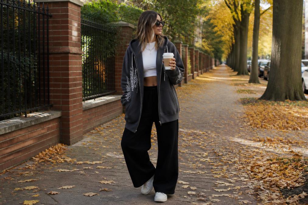 A woman layering a longline oversized graphic hoodie over a cropped top with wide-leg pants and chunky sneakers, shot outdoors in natural autumn light with fallen leaves scattered on the pavement behind her