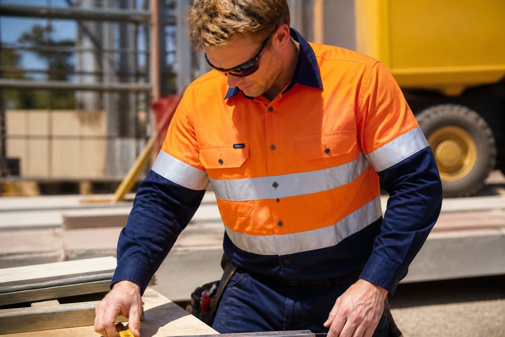 Field crew worker wearing bright orange hi-vis work shirt with reflective tape, actively working on a construction site in strong daylight, showing comfort and mobility while handling tools
