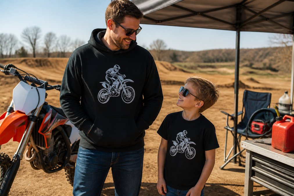 A father and young child wearing matching motocross-themed hoodies and tee shirts, standing near a dirt bike at a motocross track on a sunny day, smiling and relaxed in the pit area