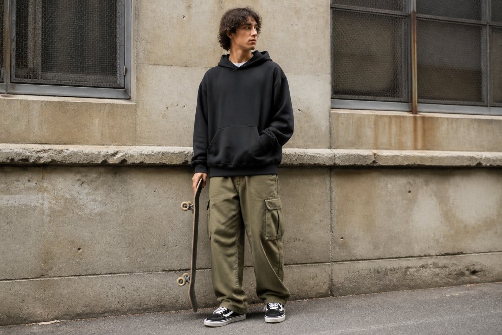 Skateboarder wearing an oversized hoodie and baggy cargo pants, standing casually with skateboard under arm in an urban street setting with concrete and graffiti walls