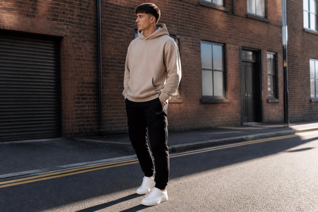 Young man wearing a sand-coloured oversized hoodie with dropped shoulders, paired with tapered black cargo pants and white trainers, standing in an urban street setting with natural afternoon light casting shadows on brick buildings