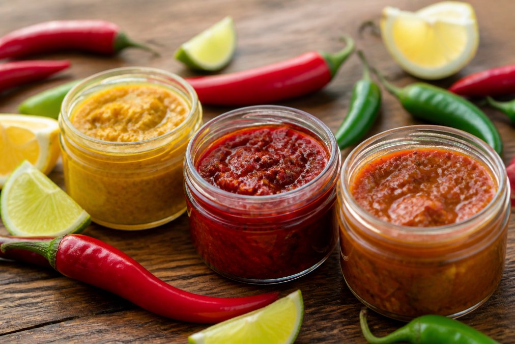 Trois petits bocaux en verre de pâte de piments artisanale disposés sur une surface en bois rustique, entourés de piments frais et de quartiers d'agrumes, sous une lumière naturelle chaude
