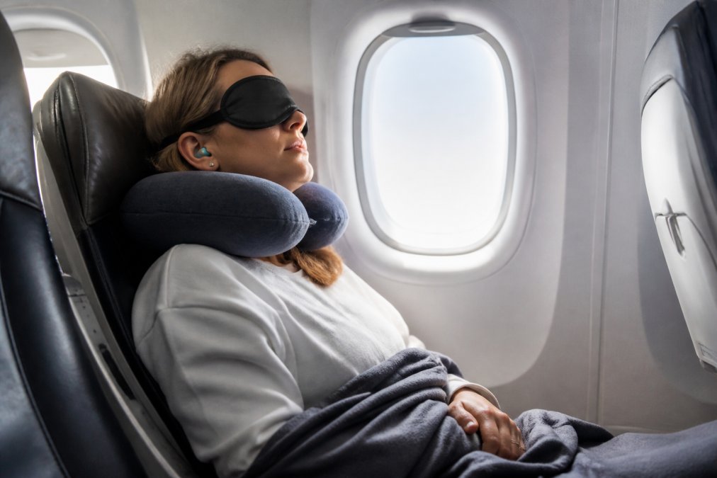 Passager confortablement installé en avion avec un coussin cervical mémoire de forme, portant un masque de sommeil 3D occultant et des bouchons d'oreilles, reposant paisiblement contre le hublot avec une couverture légère