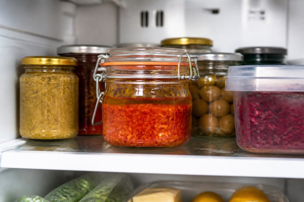 Pot de verre hermétique rempli de harissa artisanale rouge vif, recouvert d'une fine couche d'huile d'olive brillante, placé sur une étagère de réfrigérateur entre des bocaux d'autres condiments, lumière naturelle douce