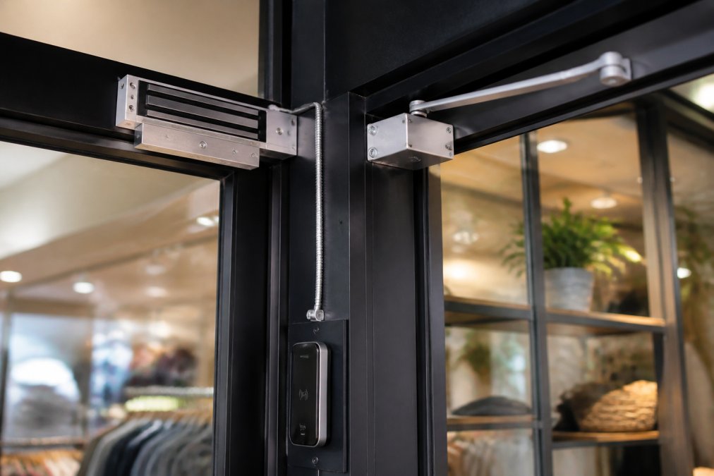 Close-up of door controller hardware with magnetic lock and access control panel mounted on a retail storefront entrance, showing the electronic components and wiring in a professional installation