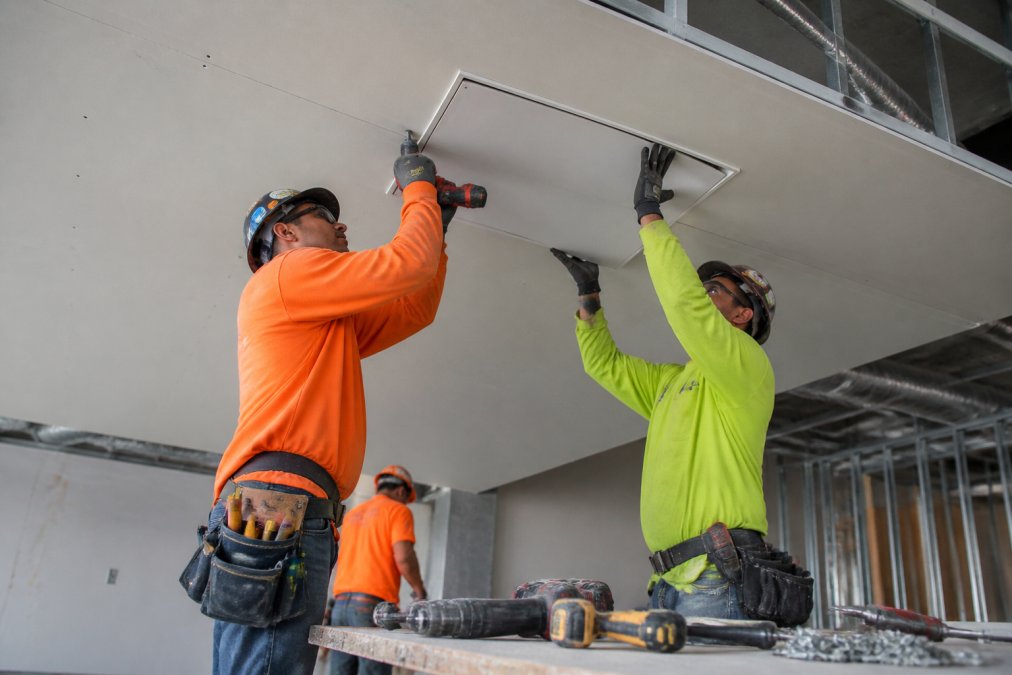 A small crew of construction workers installing a flush access door panel into a finished drywall ceiling in a commercial building, tools and hardware laid out nearby
