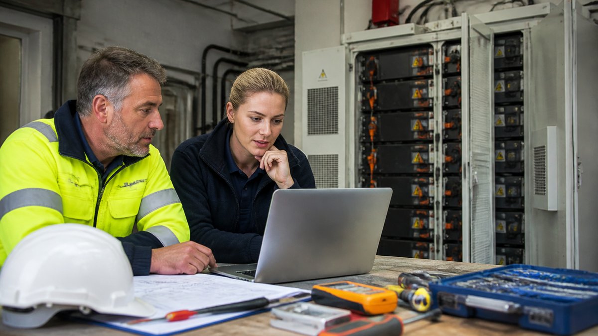 An operations manager and an energy consultant reviewing a spreadsheet of electricity bills and payback figures on a laptop in a plant room, with a battery storage cabinet visible in the background