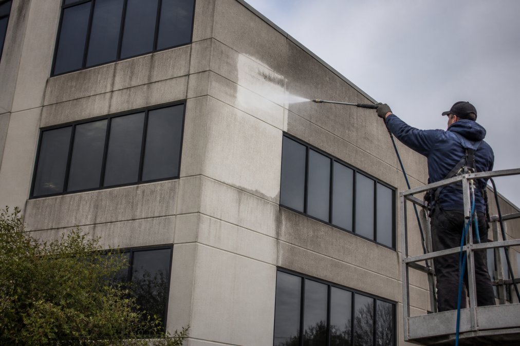 Professional pressure washing technician in safety gear using a soft wash system on a multi-story commercial building facade, with a visible clean streak contrasting against the dirty surface, overcast daylight