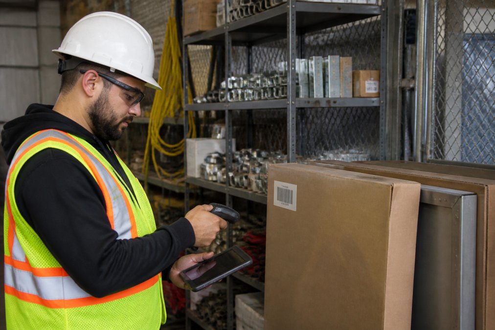 A construction site supervisor in a hard hat and safety vest using a tablet to scan a barcode on a boxed fire-rated access door stored in a secured site lockup, with organized shelving of construction hardware behind him