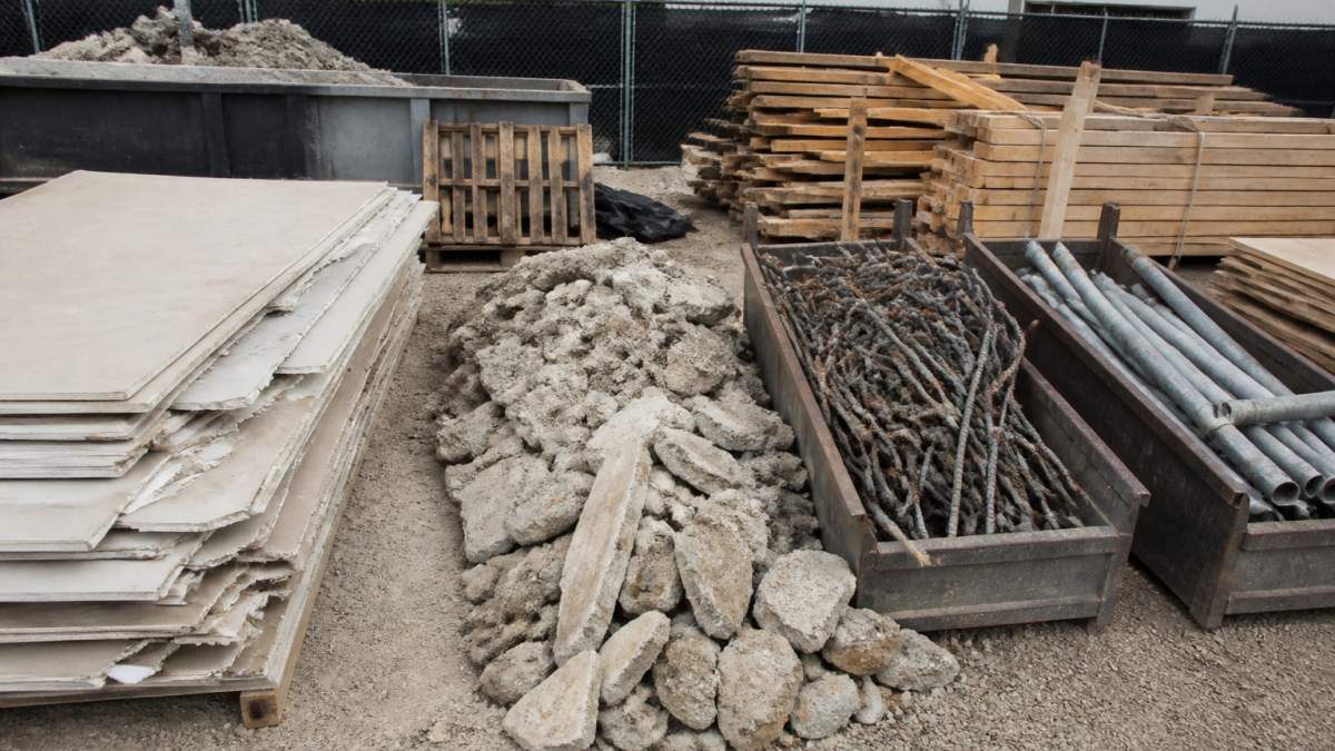 Job site showing mixed construction debris including drywall sheets stacked separately, concrete chunks piled together, scrap metal rebar and pipes sorted in bins, and wood framing materials bundled, with clear separation between material types