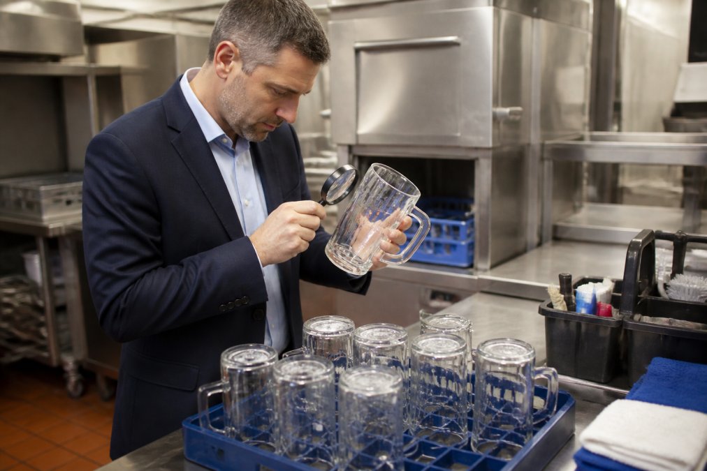 Hospitality venue manager in professional attire carefully inspecting a set of branded beer steins fresh from a commercial dishwasher, examining the etched logos with a magnifying glass and checking for cloudiness, hazing, or surface damage under bright kitchen lighting