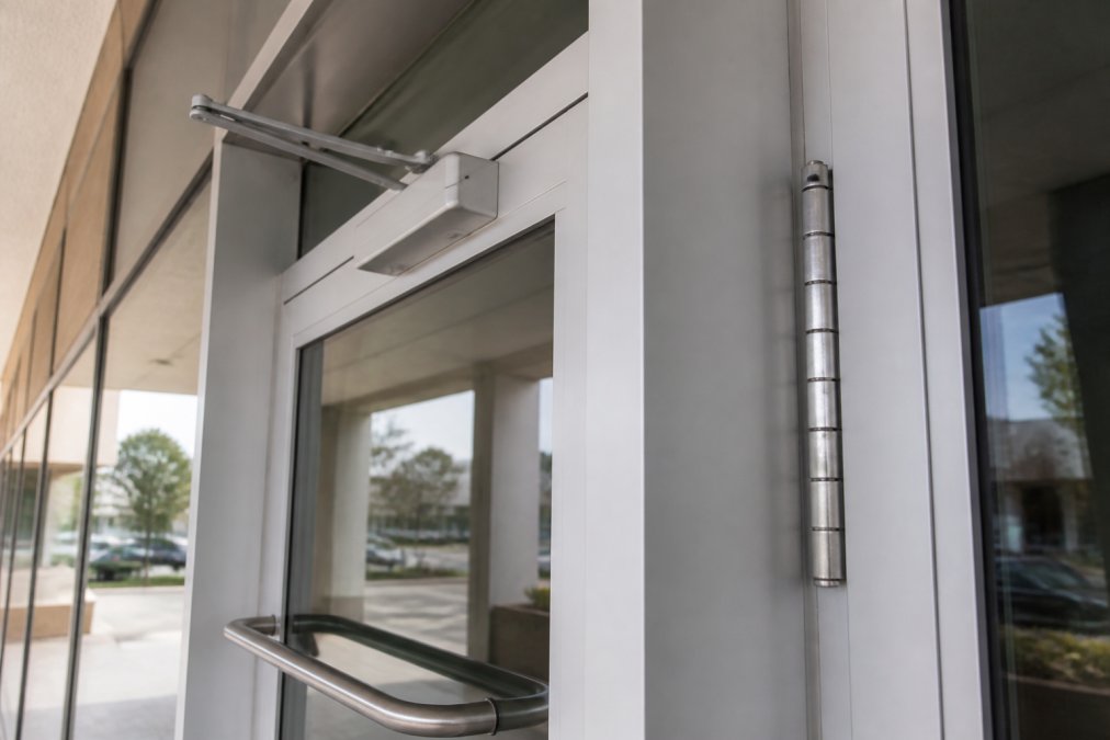 Close-up of a commercial storefront door with a heavy-duty lever handle, door closer arm, and continuous hinge visible on an aluminum frame in a well-lit commercial building entrance