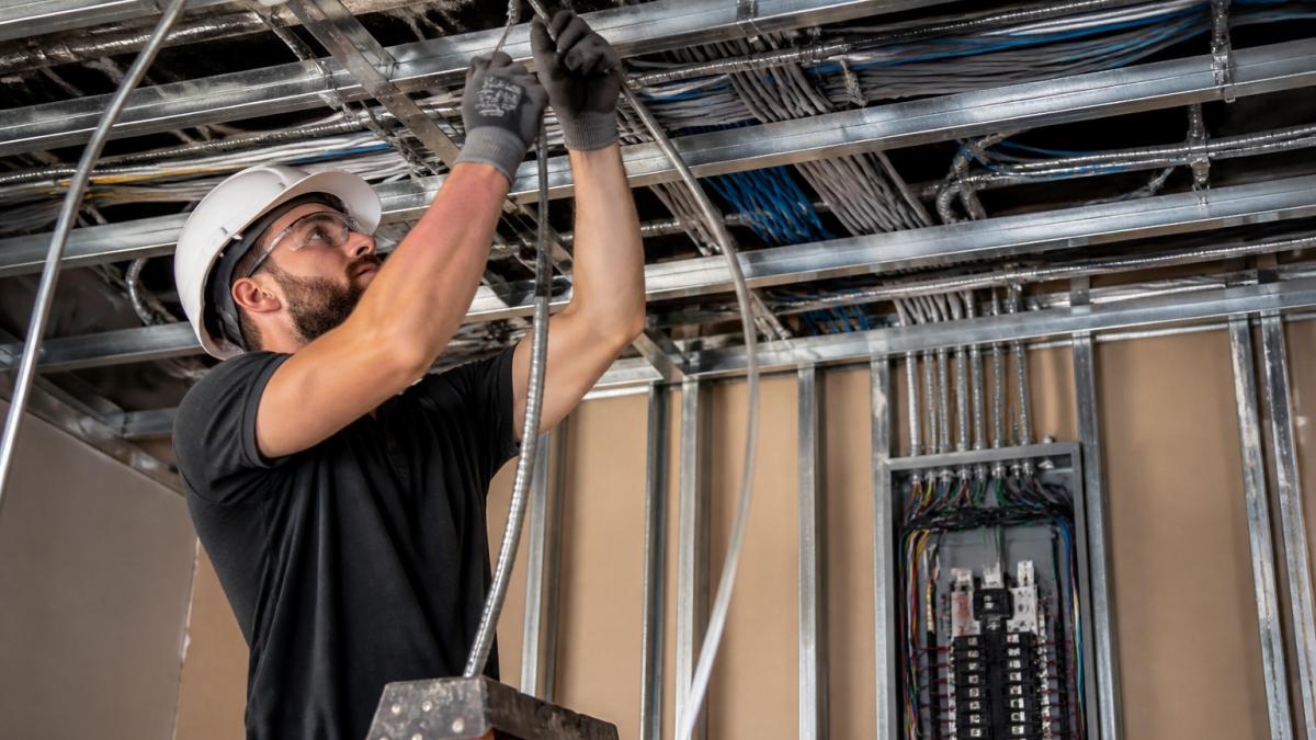 Professional electrician installing conduit and pulling wire through open ceiling space of commercial office building, with electrical panel visible in background and organized cable management