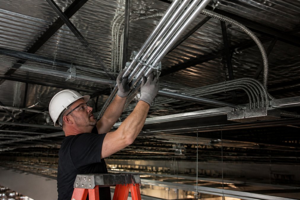 Professional electrician installing EMT conduit and MC cable in commercial office ceiling space with proper routing, labeling, and support brackets visible under bright work lighting