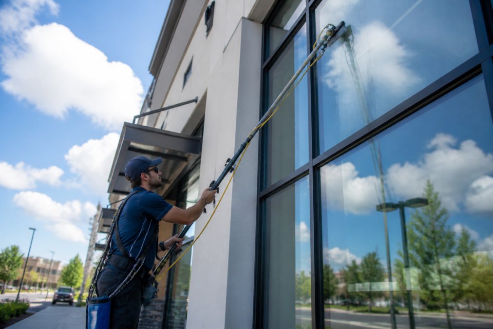 Professional window cleaner using water-fed pole system on multi-story storefront, wearing safety harness and professional uniform, streak-free glass visible in afternoon sunlight