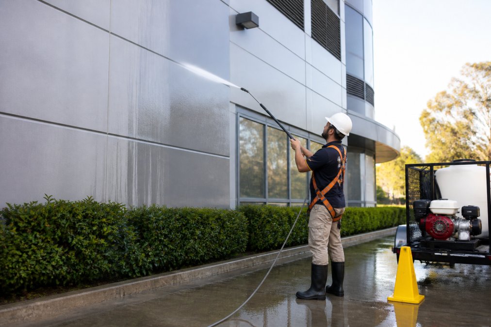 Professional demonstrating commercial exterior cleaning technique on modern commercial building facade with safety harness and high-pressure equipment, morning natural lighting
