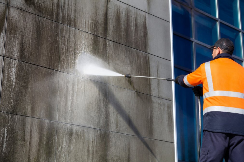 A professional in high-visibility safety gear operating a high-pressure cleaning lance on a large commercial building facade, water spray fanning out against a dark-stained concrete wall in bright midday sunlight