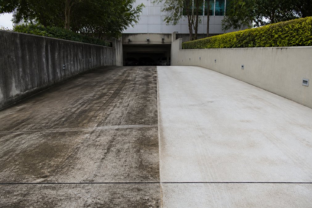 A commercial car park entrance showing a sharp contrast between the left half covered in dark algae staining and tyre marks and the right half freshly pressure-washed to a clean, bright concrete finish, under flat overcast light